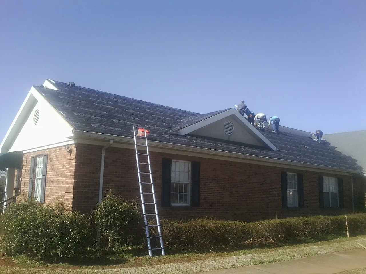 Crew tearing shingles off a brick ranch roof with a ladder set — Metal Roof Repair work by our crew in Rush